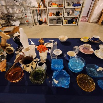 Assorted vintage glassware and ceramics, including colorful hobnail bowls, teacups, and decorative figurines displayed on a blue table.
