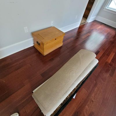 Small light wood storage chest with metal side handles, shown alongside a rolled-up tan area rug on a hardwood floor.