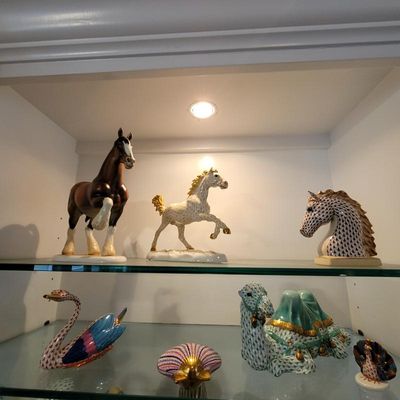Collection of fine porcelain figurines displayed on glass shelves, including Herend-style fishnet patterned animals such as a horse bust,...