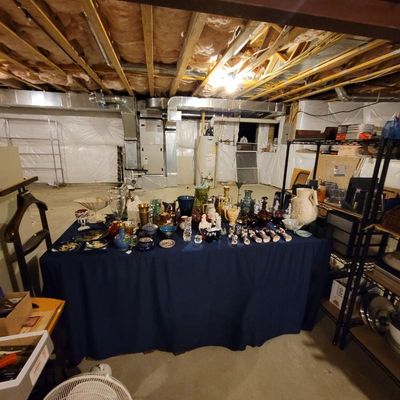 Assorted glassware, ceramics, and decorative collectibles displayed on a blue tablecloth in an unfinished basement setting.