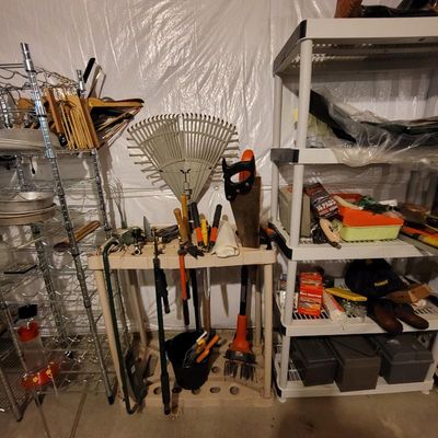 Assorted garden and hand tools, including a leaf rake, hand saw, and loppers, organized on a plastic utility cart alongside shelving...