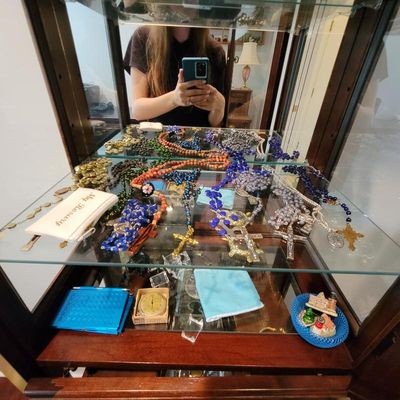 Collection of vintage rosaries in various colors and materials, displayed on a glass shelf inside a wooden curio cabinet.