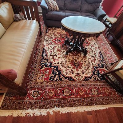 Large Persian-style area rug with intricate red, cream, and navy blue patterns and fringed edges, shown under a marble-top parlor table.