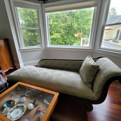 Antique-style chaise lounge featuring a curved wooden frame and elegant sage green and gold damask upholstery, positioned in front of a...
