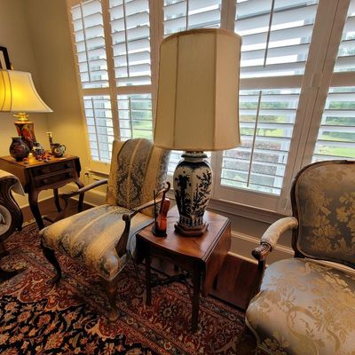 Chinoiserie blue and white ceramic table lamp with a tall fabric shade, displayed on a wooden side table between two upholstered armchairs.