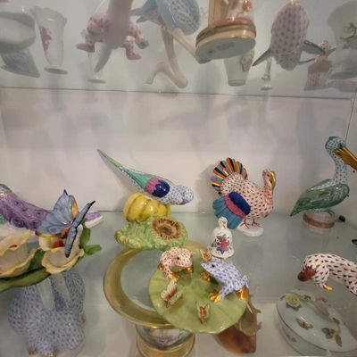 Collection of Herend porcelain figurines, including a turkey, budgerigar, frogs on a lily pad, and a pelican, displayed on glass shelves.