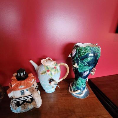 Collection of decorative ceramic items including a lighthouse teapot, a winter-themed teapot with a bird detail, and a majolica-style...