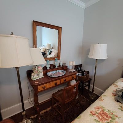 Antique wooden vanity desk with matching mirror, cane-back chair, and brass floor lamps.