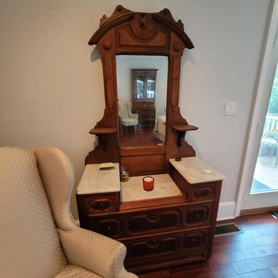 Antique Victorian walnut vanity with a tall carved mirror frame, small side shelves, and inset marble tops over a multi-drawer base.