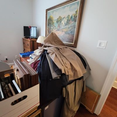 Assorted outerwear, including trench coats and jackets, piled on a table beneath a large framed landscape painting.