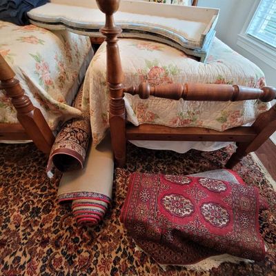 Assorted traditional patterned area rugs, shown rolled and folded alongside a pair of wooden four-poster twin beds with floral quilts.