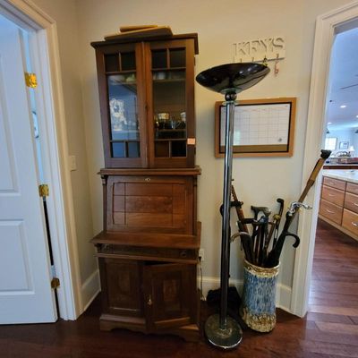 Antique wooden secretary desk with a glass-front hutch top, shown alongside a chrome torchiere floor lamp and a ceramic umbrella stand...