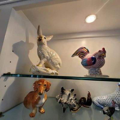 Collection of fine porcelain figurines, including a Herend-style fishnet rabbit and rooster, displayed on glass shelves.