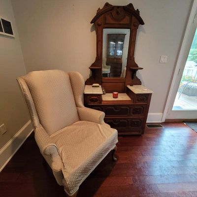 Antique Victorian marble-top dresser with an ornate wooden mirror frame, paired with a beige upholstered wingback armchair.