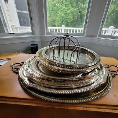 Stack of vintage silver-plated serving trays in various sizes and designs, topped with a wire toast rack.