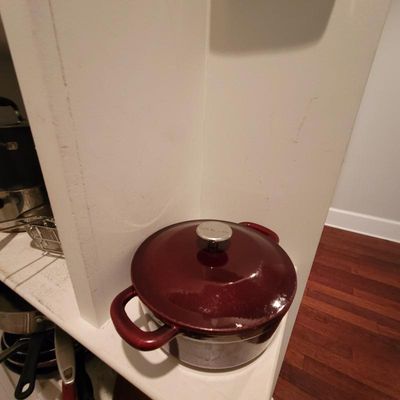 Enameled cast iron Dutch oven in a deep burgundy red with a silver-tone lid knob, shown sitting on a white shelf.