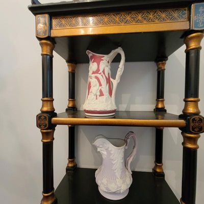 Two vintage relief-molded ceramic pitchers, one in pink and white and the other in lavender and white, displayed on an ornate black and...