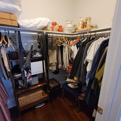 Walk-in closet containing various men's clothing, belts, a wooden shelving unit, and decorative glassware on the top shelf.