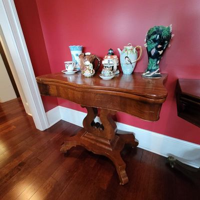 Antique Empire-style mahogany card table with a lyre-shaped pedestal base, displaying an assortment of vintage ceramic teapots, pitchers,...