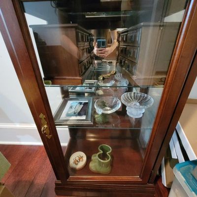 Vintage wooden curio cabinet with glass shelves and a mirrored back, displaying small collectibles, glassware, and framed photos.