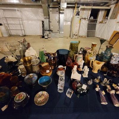 A large collection of vintage glassware, ceramics, and decorative figurines displayed on a table in a basement setting.