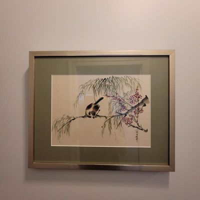 Framed East Asian ink and wash painting depicting a bird perched on a branch with pink blossoms, presented in a silver-toned frame with a...