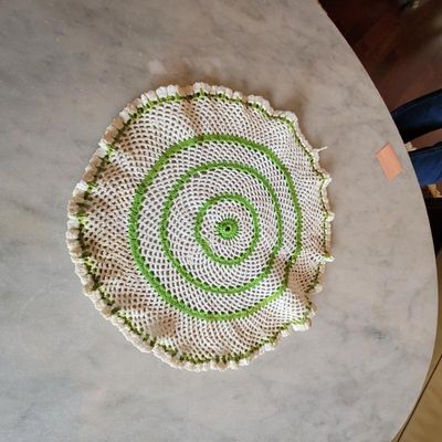 Vintage handmade crochet doily featuring concentric green and white circular patterns with a ruffled edge, displayed on a marble tabletop.