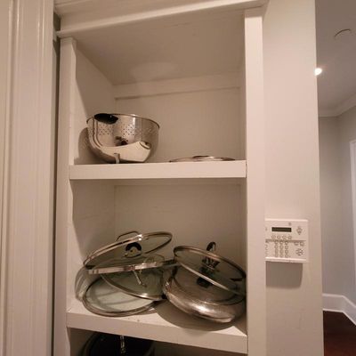 Assorted stainless steel cookware lids and a metal colander stored on white wooden shelves.