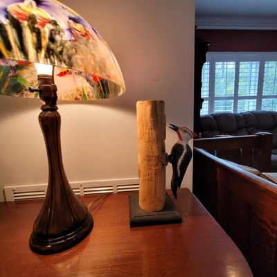 Vintage table lamp with a colorful floral glass shade alongside a hand-carved wooden woodpecker sculpture on a tree trunk base.