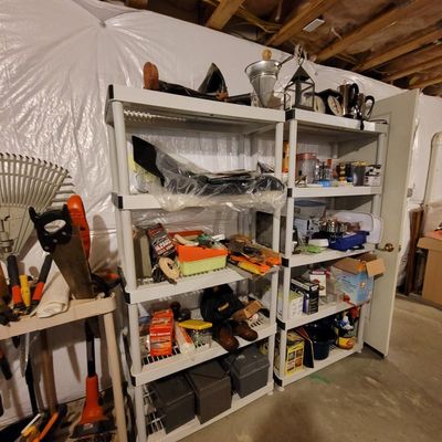 Two white plastic five-tier shelving units filled with various household items, tools, and storage bins in a basement setting.