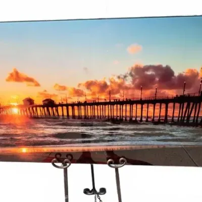Frameless acrylic or metal print depicting a sunset over a coastal pier, displayed on a metal easel.