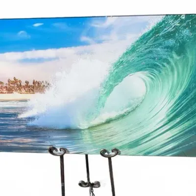 Large frameless acrylic wall art depicting a curling ocean wave crashing near a palm tree-lined beach, displayed on a black metal easel.