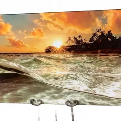 Frameless acrylic wall art depicting a tropical sunset over ocean waves and palm trees.
