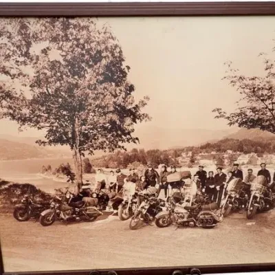Vintage sepia-toned photograph depicting a group of motorcyclists gathered with their bikes on a scenic overlook, housed in a simple dark...
