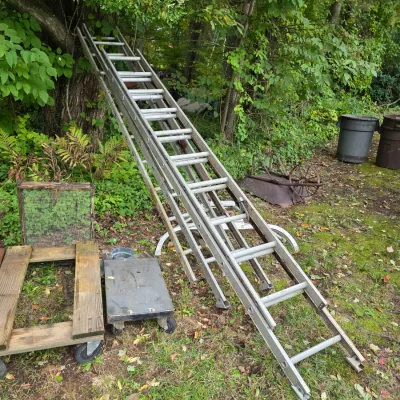 Sale Photo Thumbnail #18: Aluminum extension ladder leaning against a tree, shown alongside a wooden dolly, a metal cart, and other outdoor items.