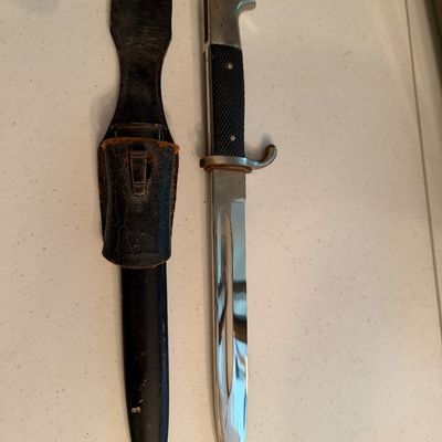 Sale Photo Thumbnail #115: German WWII-era dress bayonet featuring a polished steel blade, checkered black grips, and original metal scabbard with a leather frog.