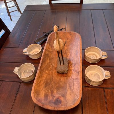 Rustic wooden dough bowl centerpiece holding a carved bird figurine, flanked by four handmade ceramic mugs on a dark wood dining table.