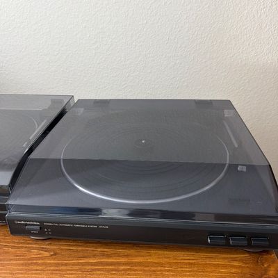 Audio-Technica AT-PL50 stereo full automatic turntable system with a tinted dust cover, resting on a wooden shelf.