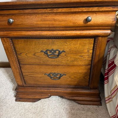 Sale Photo Thumbnail #193: Traditional oak nightstand featuring a top pull-out tray with metal knobs and two lower drawers with ornate metal bail pulls.