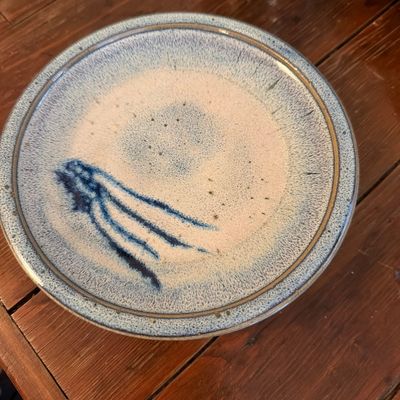 Studio art pottery plate featuring a speckled beige and blue glaze with a stylized blue abstract design, resting on a wooden surface.
