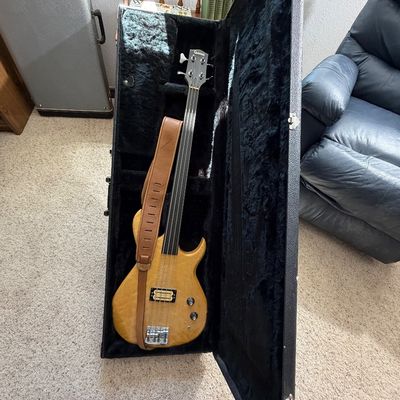 Vintage Carvin fretless electric bass guitar with a natural wood finish, resting inside its plush-lined black hard-shell case alongside a...