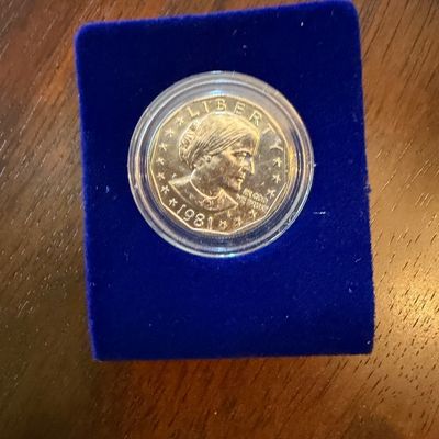 Sale Photo Thumbnail #210: 1981 Susan B. Anthony dollar coin, housed in a protective plastic capsule and mounted on a blue velvet display stand.