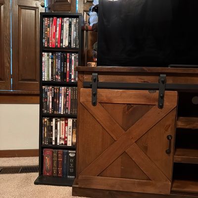 Sale Photo Thumbnail #135: Rustic wooden media console featuring a sliding barn-style door with black metal hardware, shown next to a black five-shelf media tower filled with DVDs.