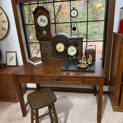 Mission-style oak writing desk with a single drawer, paired with a matching wooden stool and decorated with an assortment of vintage clocks.