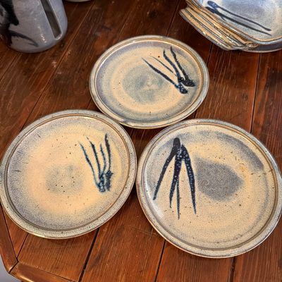 Three studio art pottery plates featuring a speckled beige and blue glaze with abstract dark blue brushstrokes, displayed on a wooden...