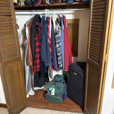 Sale Photo Thumbnail #196: Assorted vintage and modern clothing, handbags, and luggage, including a black rolling suitcase and green duffel bags, organized inside a closet with louvered doors.