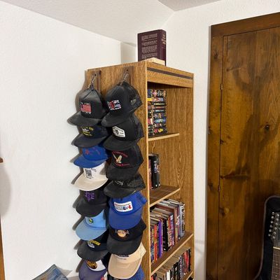 Sale Photo Thumbnail #186: Tall oak-finish bookcase filled with books, featuring two hanging vertical cap organizers displaying a collection of vintage trucker hats and baseball caps.