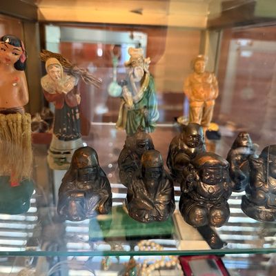 Sale Photo Thumbnail #113: Collection of vintage miniature figurines displayed inside a glass cabinet, including a Hawaiian hula girl nodder, painted Asian sage figures, and small dark metal deities.