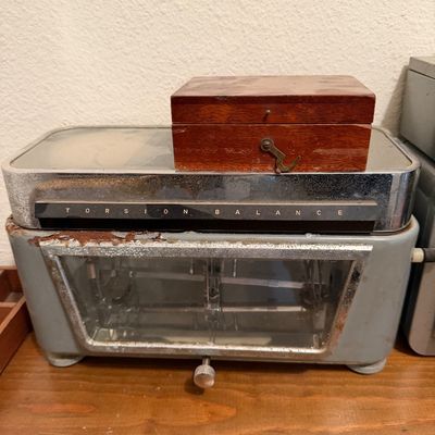 Vintage Torsion Balance laboratory scale with a grey metal housing and glass viewing window, topped with a small wooden weight box.