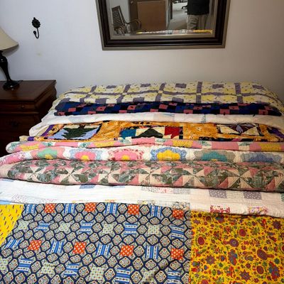 A collection of vintage handmade quilts in various colorful patchwork, floral, and geometric patterns, displayed layered on a bed.